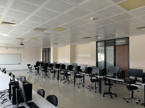 L110 Bilgisayar Laboratuvarında duvar boyunca yerleştirilmiş bilgisayarlar, çalışma masaları ve koltuklar.
