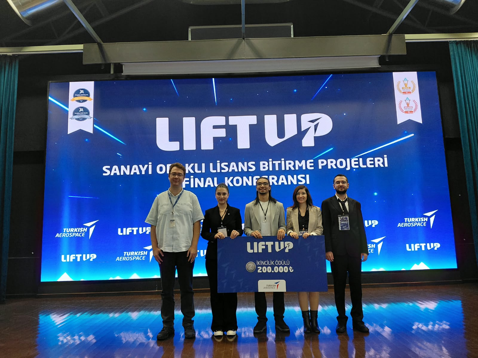 Öğrencilerimiz Bekir Berk YILDIRIM, Yaren DİNÇ ve Mert ÖZDEMİR, akademik danışmanları Doç. Dr. Hilal ARSLAN ve TUSAŞ sanayi danışmanı Abdullah Nuri SOMUNCUOĞLU ile ödül töreninde sahnede ödüllerini sergiliyorlar.