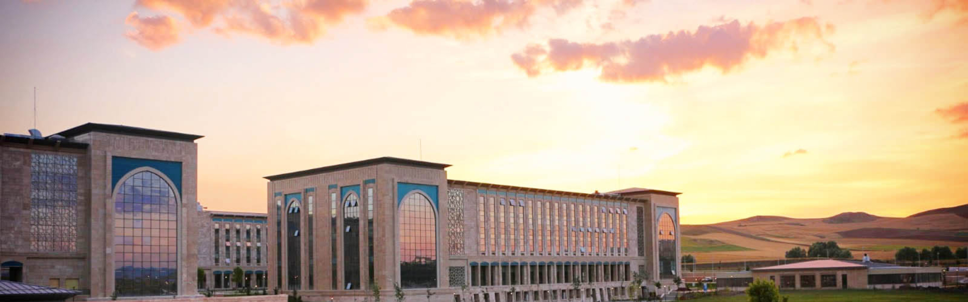 AYBÜ Esenboğa Külliyesi binalarının gün batımı saatlerinde çekilmiş genel görünümü; ön planda yeşil peyzaj alanları ve arkada geniş gökyüzü manzarası.