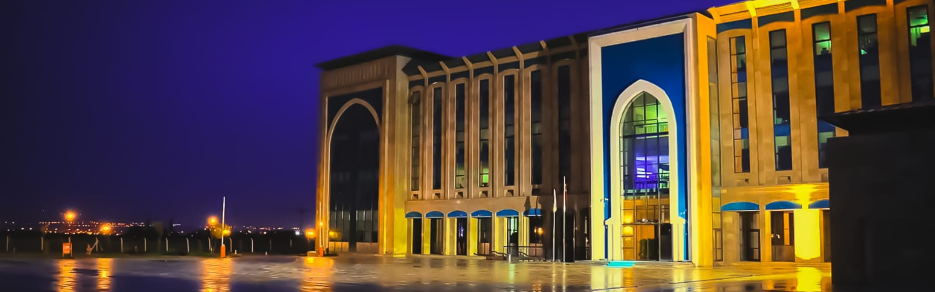 Ankara Yıldırım Beyazıt Üniversitesi Esenboğa Külliyesi'nin gece görünümü. Işıklandırılmış modern Selçuklu mimarisine sahip rektörlük binası ve önündeki geniş meydan karanlık gökyüzü altında parlıyor.