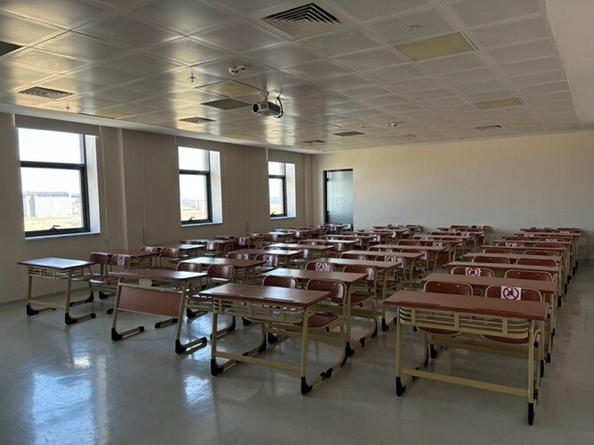 L115 Dersliğinde sıralı öğrenci masaları, sandalyeler ve tavana monte projeksiyon cihazı.